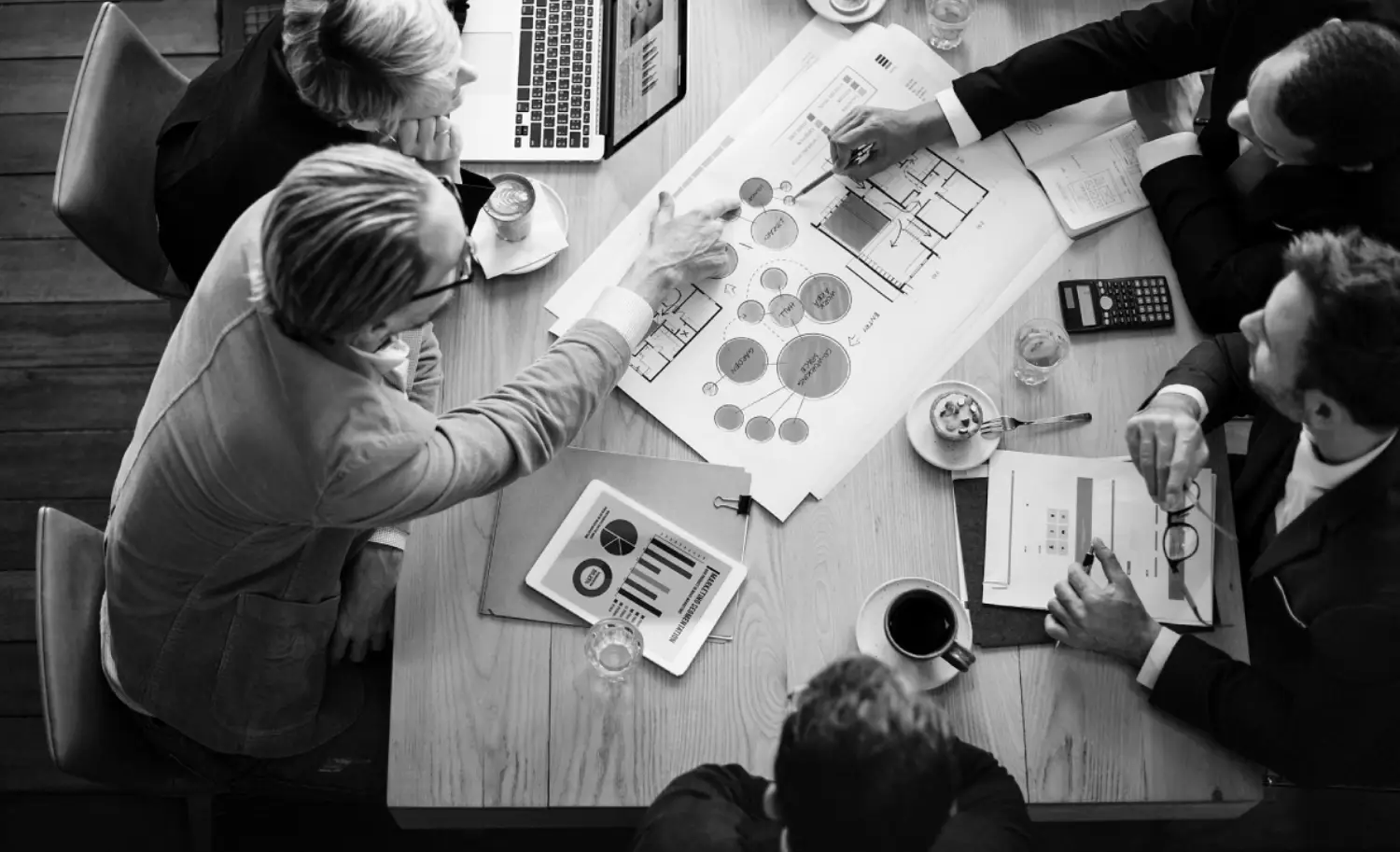 A group of professionals collaborates around a table, analyzing charts and plans, with laptops and drinks present